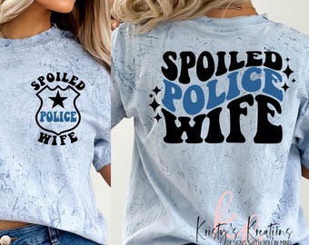 May include: A light blue t-shirt with the text "Spoiled Police Wife" in black and blue retro font. The shirt has a distressed look and is shown on a model wearing blue jeans.