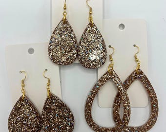 May include: Three pairs of teardrop-shaped earrings featuring a dense, multi-coloured glitter finish. Each pair is suspended from gold-toned metal fishhook wires and displayed on individual white rectangular cards with a circular hole at the top.
