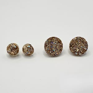 May include: Two pairs of circular stud earrings featuring a textured, glittery gold surface. The set includes one pair of smaller circular studs and one pair of larger circular studs, both displaying a reflective, multi-faceted finish.