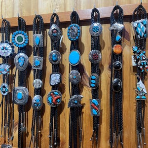 Può includere: Una collezione di cravatte bolo con vari pendenti decorativi. I pendenti presentano turchesi, argento e altre pietre e disegni colorati. Le cravatte sono appese su uno sfondo di legno, mostrando la varietà di stili.