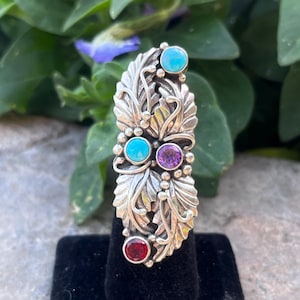May include: A silver ring with an elaborate leaf pattern. The ring is adorned with four gemstones in turquoise, amethyst, and garnet. The ring is displayed on a black velvet surface, with green foliage in the background.