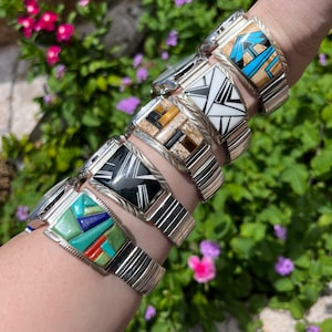 Puede incluir: Una colección de seis relojes plateados con incrustaciones de piedras de diferentes colores en la esfera. Los relojes tienen correas de metal plateado.