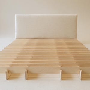 May include: A modern bed frame with a light-colored wooden slat base and a rectangular, off-white upholstered headboard. The bed frame is made of natural wood and is designed for a minimalist bedroom.
