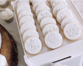 May include: A white tray holds numerous round, white cookies, each with a unique embossed design. Designs include floral patterns, the words "we do", and interlocking rings. Some cookies are embossed with "Mr & Mrs".