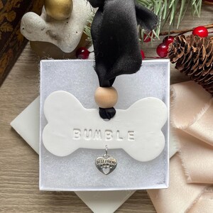 May include: White ceramic dog bone ornament with the name "Bumble" inscribed on it. The ornament is hanging from a black ribbon with a wooden bead. A silver charm with the words "Best Friend" is attached to the ornament.
