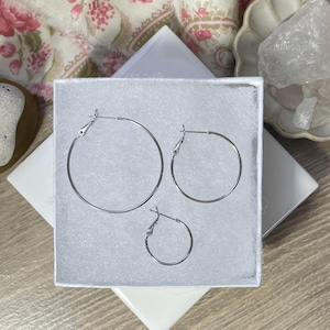 May include: Three silver hoop earrings of different sizes in a white box. The earrings are all circular and have a simple design.