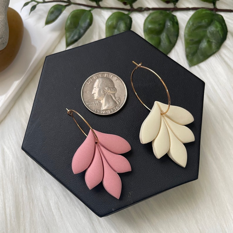 Pode incluir: Um par de brincos de argola dourados com pingentes em forma de flor rosa e branca. Os brincos s&atilde;o exibidos em uma superf&iacute;cie hexagonal preta.