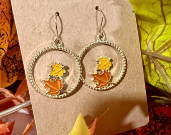 May include: Gold-tone hoop earrings featuring a fall leaf design. Each earring has a circular frame with a textured edge, and inside, there are two maple leaves in shades of yellow and orange. The earrings are displayed on a brown card.