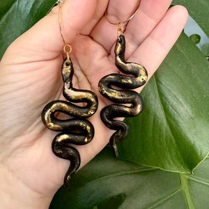 May include: A pair of black and gold snake earrings. The snakes are made of polymer clay and have a textured finish. The earrings are hanging from gold hoops.