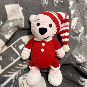 Könnte beinhalten: Ein roter gehäkelter Teddybär, der eine weiß-rot gestreifte Mütze trägt. Der Bär hat ein rosafarbenes Gesicht und schwarze Augen. Der Bär sitzt auf einer grau-weißen Decke.