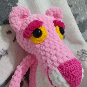 May include: Pink crocheted plush toy shaped like the Pink Panther cartoon character. The toy has black eyes with yellow pupils and a pink nose.