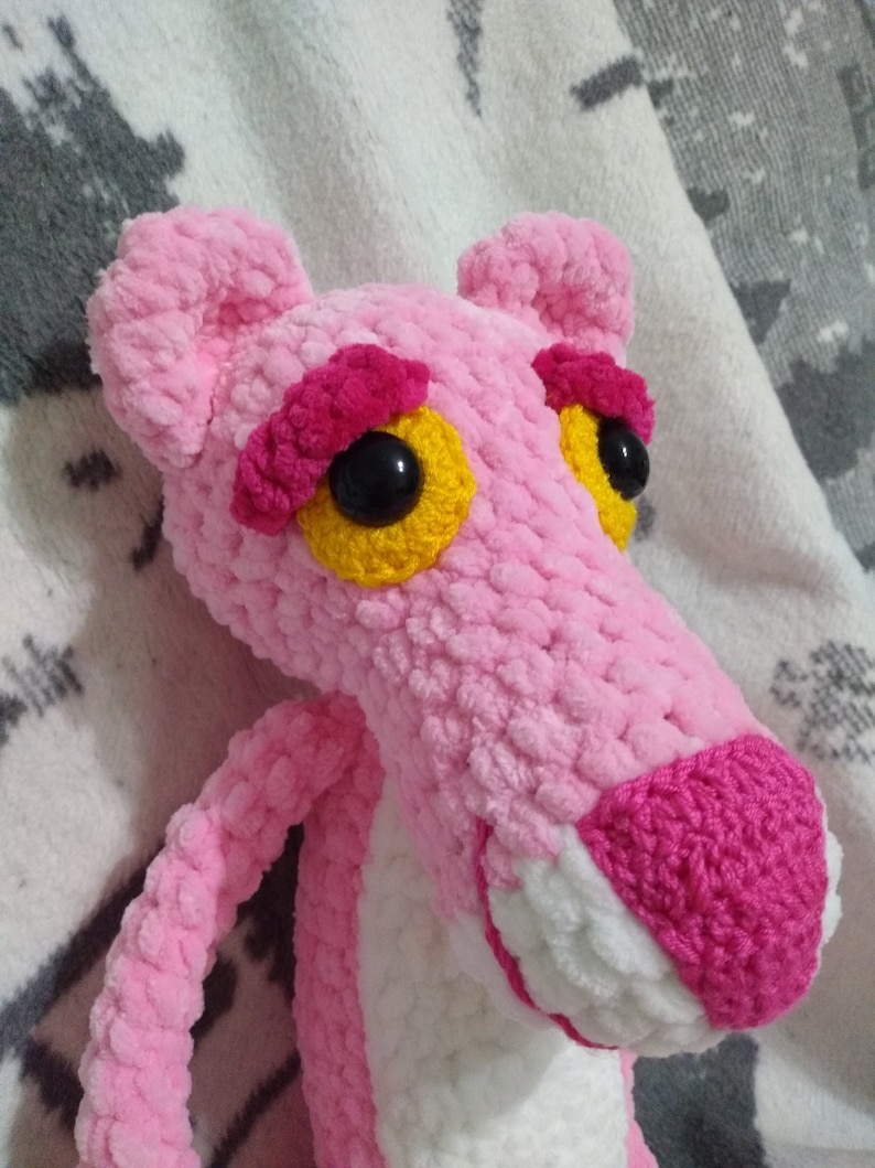 May include: Pink crocheted plush toy shaped like the Pink Panther cartoon character. The toy has black eyes with yellow pupils and a pink nose.