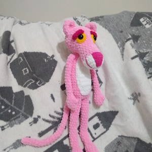 May include: A pink crocheted plush toy shaped like the Pink Panther cartoon character. The toy has a white chest and yellow eyes.
