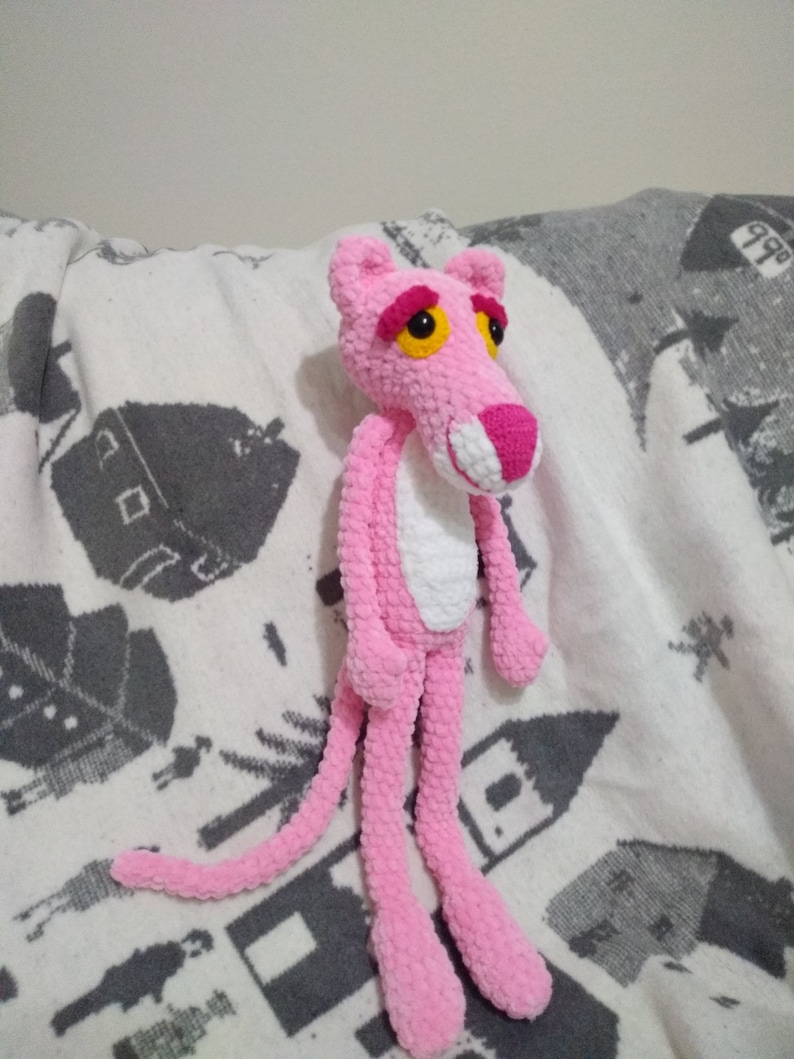 May include: A pink crocheted plush toy shaped like the Pink Panther cartoon character. The toy has a white chest and yellow eyes.