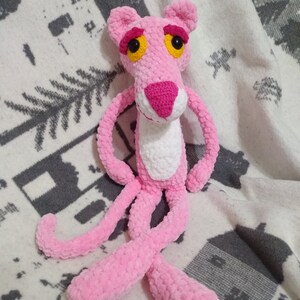 May include: Pink crocheted plush toy of the Pink Panther cartoon character. The toy has a white belly and large yellow eyes.