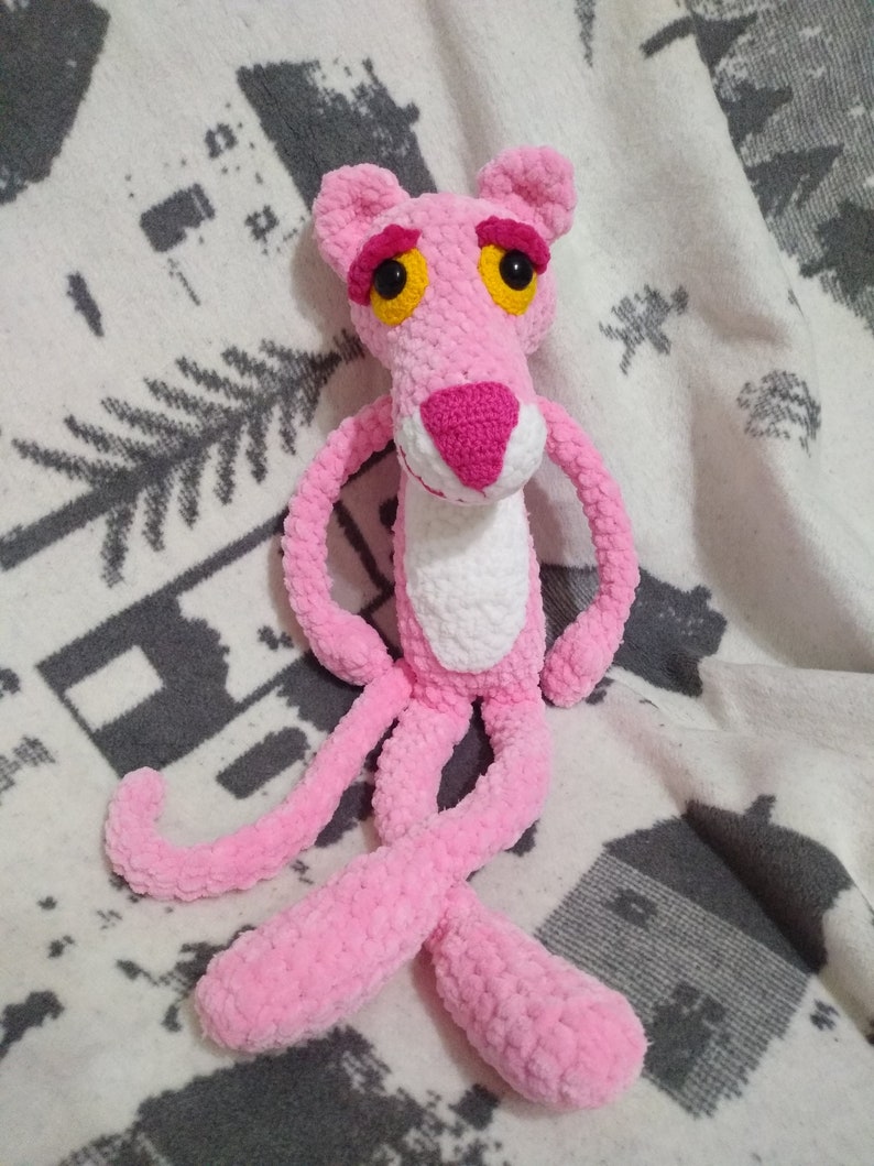 May include: Pink crocheted plush toy of the Pink Panther cartoon character. The toy has a white belly and large yellow eyes.