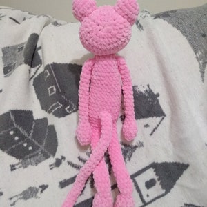 May include: A pink crocheted plush toy shaped like a monkey with long arms and legs. The toy is sitting on a white and gray blanket.