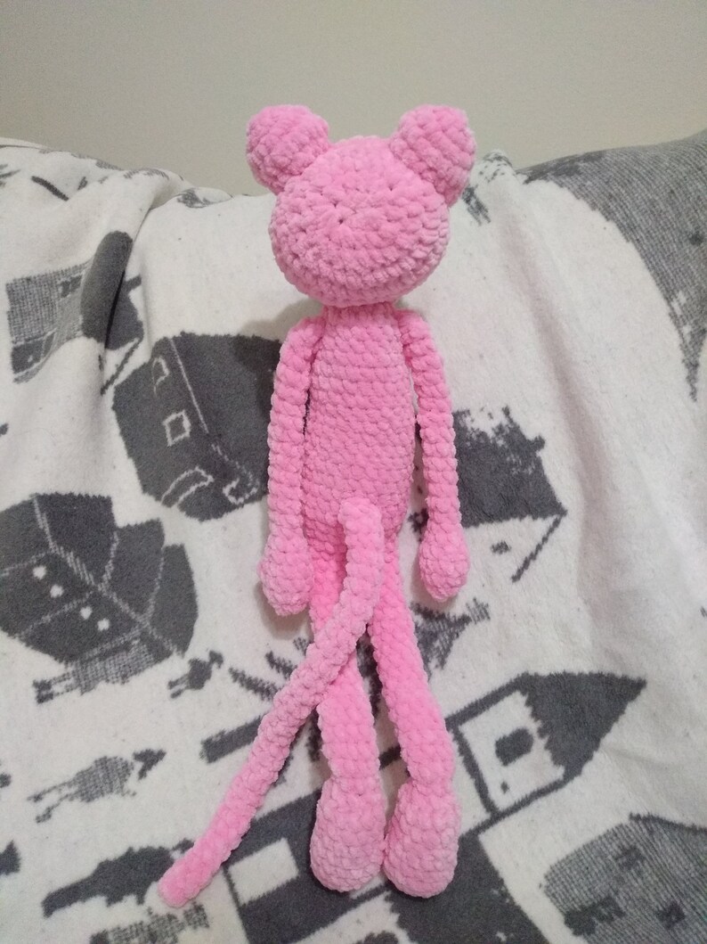 May include: A pink crocheted plush toy shaped like a monkey with long arms and legs. The toy is sitting on a white and gray blanket.