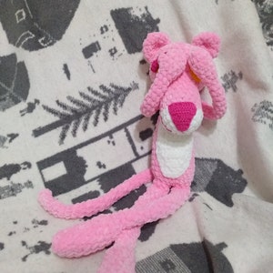 May include: Pink crocheted plush toy shaped like a panther with its eyes covered by its paws. The toy is sitting on a white and gray patterned blanket.