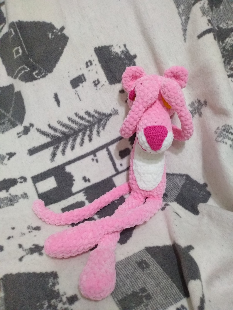 May include: Pink crocheted plush toy shaped like a panther with its eyes covered by its paws. The toy is sitting on a white and gray patterned blanket.