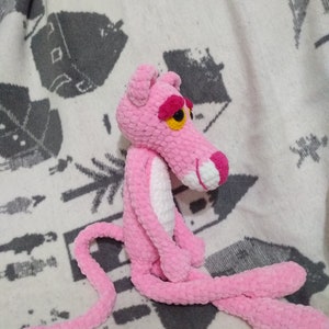 May include: A pink crocheted plush toy shaped like the Pink Panther. The toy has a white belly and a long pink tail.