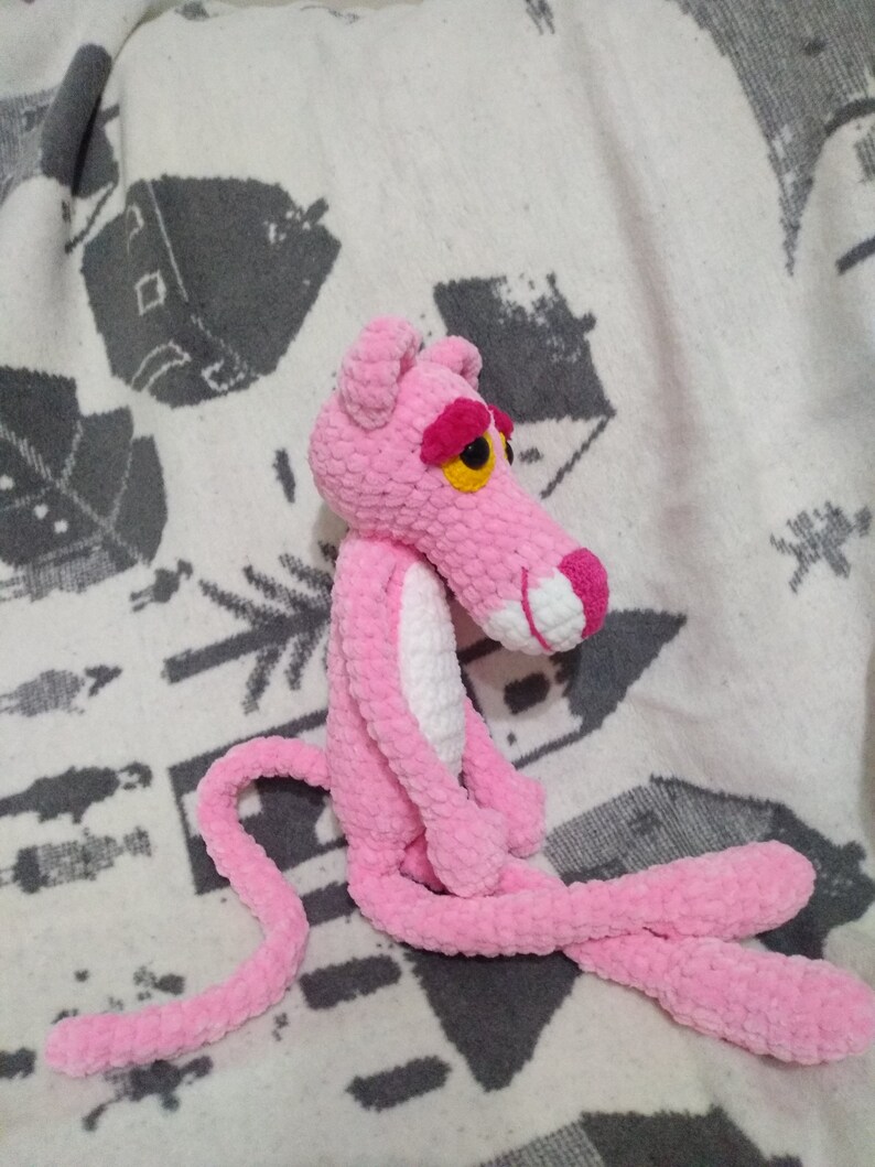 May include: A pink crocheted plush toy shaped like the Pink Panther. The toy has a white belly and a long pink tail.