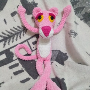 May include: Pink crocheted plush toy of the Pink Panther cartoon character. The toy has a white belly and yellow eyes.