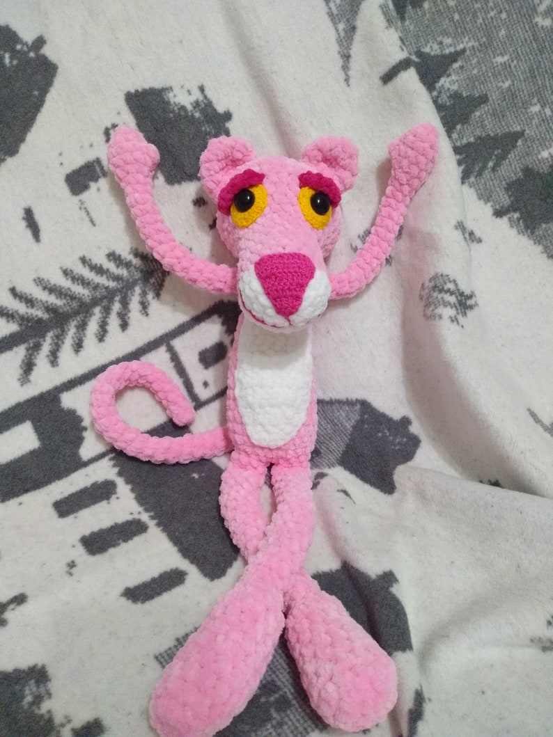 May include: Pink crocheted plush toy of the Pink Panther cartoon character. The toy has a white belly and yellow eyes.
