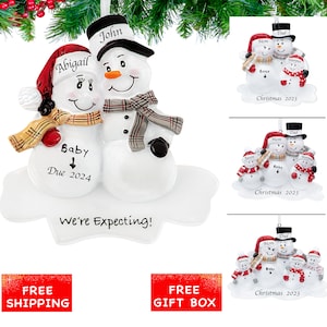 May include: A white snowman ornament with a red hat and scarf, holding a smaller snowman with a red hat and scarf. The larger snowman has the text "John" on its hat and the smaller snowman has the text "Abigail" on its hat. The smaller snowman has a sign that says "Baby Due 2024". The ornament also has the text "We're Expecting!" on it.