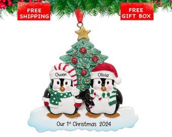 May include: A Christmas tree ornament featuring two penguins standing on a snowy base. The left penguin wears a red and white striped hat and green scarf with white dots, labeled Owen. The right penguin wears a red hat and green striped scarf, labeled Olivia. Text reads Our 1st Christmas 2024.