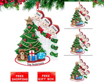 May include: A personalized Christmas ornament featuring a family of five standing around a decorated Christmas tree. The ornament is white with a green tree and red and gold decorations. The family members are all wearing Santa hats and have their names written on their ornaments. The ornament also includes the year 2023 and the text "The Andersons".