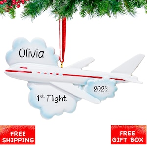 May include: White airplane ornament with red accents and the text "Olivia", "1st Flight", and "2025". The ornament is attached to a red ribbon and is surrounded by a cloud-like design.