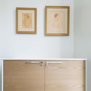 Puede incluir: Un armario blanco con dos puertas de madera y tiradores plateados. El armario está montado en una pared blanca sobre un suelo de baldosas. Dos dibujos enmarcados de mujeres cuelgan de la pared sobre el armario.