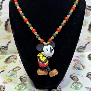 Puede incluir: Un collar con colgante de Mickey Mouse. El collar presenta un dije de Mickey Mouse con una combinaci&oacute;n de colores negro, blanco, rojo, amarillo y marr&oacute;n. El collar de cuentas tiene cuentas rojas, amarillas y negras.