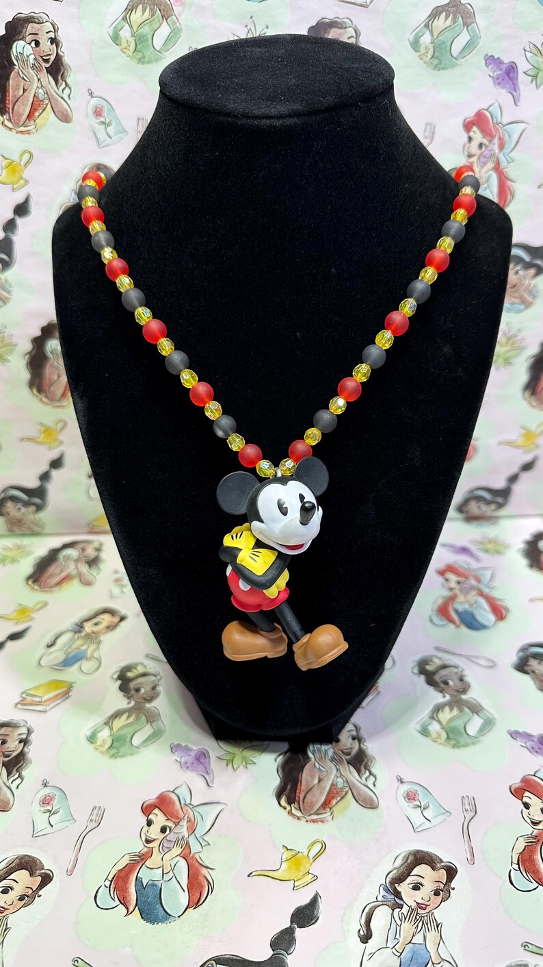 Puede incluir: Un collar con colgante de Mickey Mouse. El collar presenta un dije de Mickey Mouse con una combinaci&oacute;n de colores negro, blanco, rojo, amarillo y marr&oacute;n. El collar de cuentas tiene cuentas rojas, amarillas y negras.