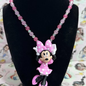 May include: A necklace featuring a Minnie Mouse pendant with a pink polka dot dress and bow, suspended from a strand of alternating light pink, dark pink, and clear translucent beads. The pendant depicts the character with white gloves.