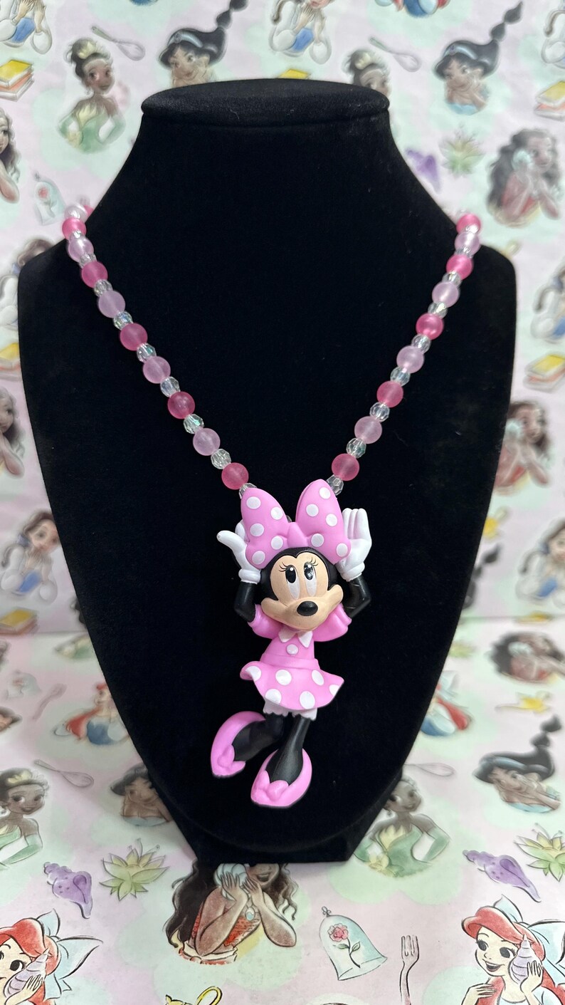 May include: A necklace featuring a Minnie Mouse pendant with a pink polka dot dress and bow, suspended from a strand of alternating light pink, dark pink, and clear translucent beads. The pendant depicts the character with white gloves.