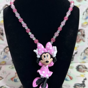 May include: A necklace featuring a Minnie Mouse pendant with a pink dress and bow with white polka dots, white gloves, and pink shoes. The chain is composed of alternating translucent clear and opaque pink spherical beads in varying shades.
