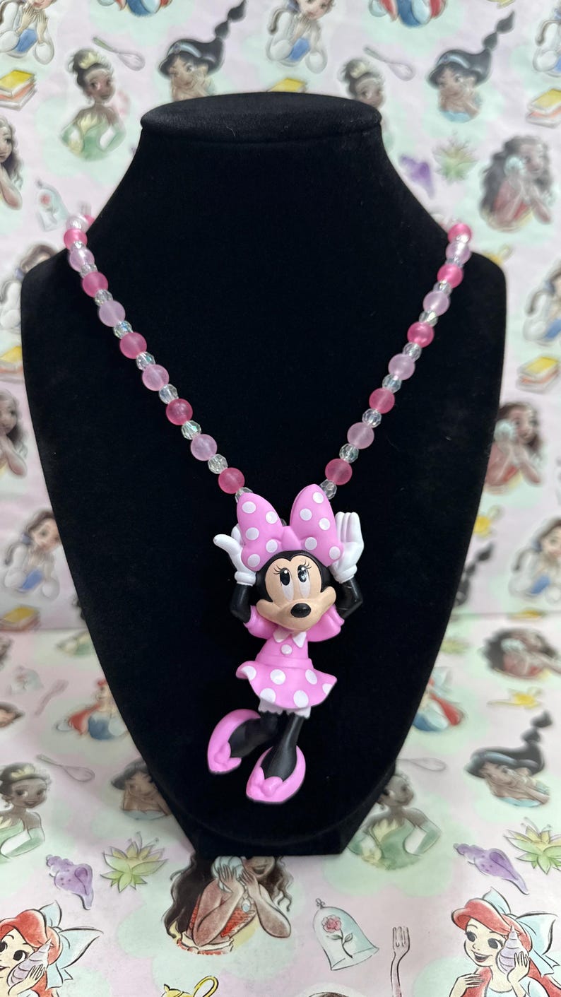 May include: A necklace featuring a Minnie Mouse pendant with a pink dress and bow with white polka dots, white gloves, and pink shoes. The chain is composed of alternating translucent clear and opaque pink spherical beads in varying shades.