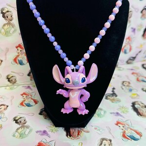 K&ouml;nnte beinhalten: Eine Halskette mit einer violetten und rosafarbenen Perlenkette und einem zentralen Anh&auml;nger der Figur Stitch. Der Anh&auml;nger ist eine hellviolette Alien-Figur mit gro&szlig;en Ohren, dunkelblauen Augen und einem hellrosa Brustfleck, die an der Kette h&auml;ngt.