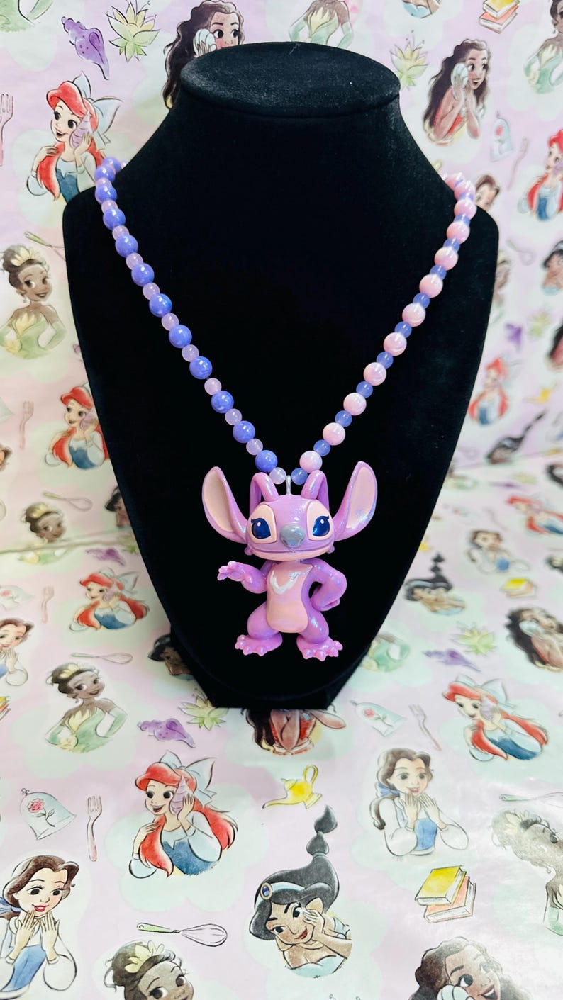 K&ouml;nnte beinhalten: Eine Halskette mit einer violetten und rosafarbenen Perlenkette und einem zentralen Anh&auml;nger der Figur Stitch. Der Anh&auml;nger ist eine hellviolette Alien-Figur mit gro&szlig;en Ohren, dunkelblauen Augen und einem hellrosa Brustfleck, die an der Kette h&auml;ngt.
