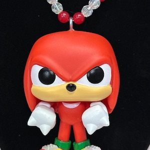 May include: A necklace featuring a red Knuckles character pendant with large white fists, a tan muzzle, and black eyes. The character wears green and yellow shoes. The necklace chain consists of alternating clear iridescent and red spherical beads.