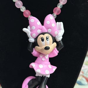 May include: A necklace featuring a Minnie Mouse pendant with a pink dress and bow with white polka dots, black limbs, and white gloves. The chain consists of alternating translucent clear and matte pink spherical beads in a repeating pattern.