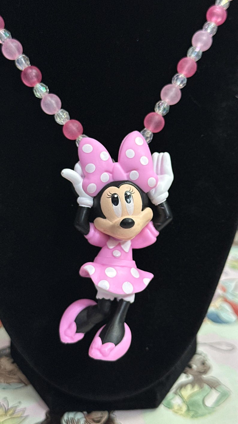 May include: A necklace featuring a Minnie Mouse pendant with a pink dress and bow with white polka dots, black limbs, and white gloves. The chain consists of alternating translucent clear and matte pink spherical beads in a repeating pattern.