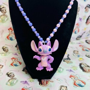 K&ouml;nnte beinhalten: Eine Halskette mit einem hellvioletten und rosafarbenen Anh&auml;nger der Figur Angel aus Lilo und Stitch, befestigt an einer Perlenkette aus abwechselnd hellvioletten und hellrosa runden Perlen, pr&auml;sentiert auf einer schwarzen Samtb&uuml;ste.