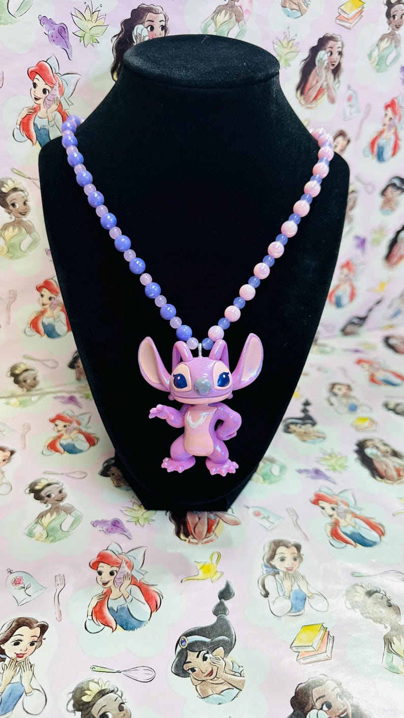 K&ouml;nnte beinhalten: Eine Halskette mit einem hellvioletten und rosafarbenen Anh&auml;nger der Figur Angel aus Lilo und Stitch, befestigt an einer Perlenkette aus abwechselnd hellvioletten und hellrosa runden Perlen, pr&auml;sentiert auf einer schwarzen Samtb&uuml;ste.