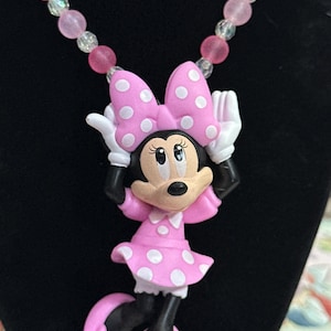 May include: A necklace featuring a Minnie Mouse pendant with a pink polka dot dress and bow, white gloves, and pink shoes. The chain consists of alternating translucent clear and matte pink spherical beads, creating a colorful and playful accessory.