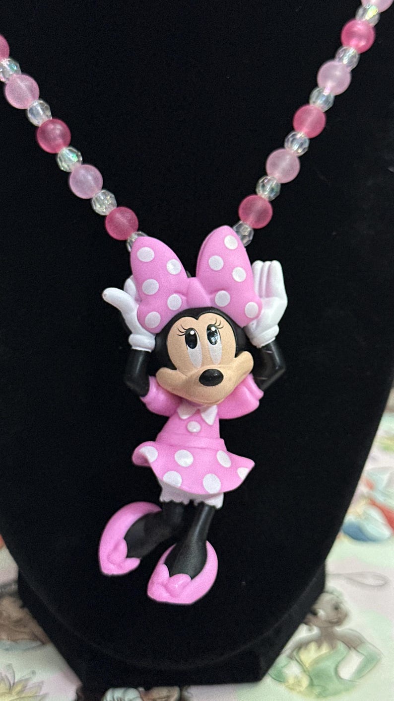 May include: A necklace featuring a Minnie Mouse pendant with a pink polka dot dress and bow, white gloves, and pink shoes. The chain consists of alternating translucent clear and matte pink spherical beads, creating a colorful and playful accessory.