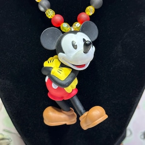 Puede incluir: Un collar con colgante de Mickey Mouse. El colgante presenta a Mickey Mouse con su atuendo cl&aacute;sico, con pantalones cortos rojos, guantes amarillos y zapatos marrones. El collar tiene una cadena de cuentas rojas, amarillas y grises. El fondo es negro.