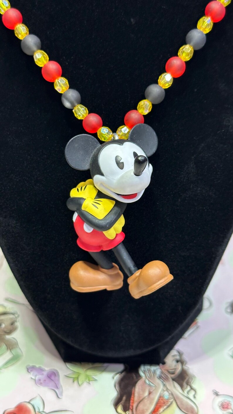 Puede incluir: Un collar con colgante de Mickey Mouse. El colgante presenta a Mickey Mouse con su atuendo cl&aacute;sico, con pantalones cortos rojos, guantes amarillos y zapatos marrones. El collar tiene una cadena de cuentas rojas, amarillas y grises. El fondo es negro.